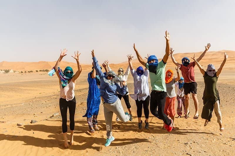 Multiple people jumping for joy in the desert, vibrantly dressed in colorful Nomadic scarfs after an adventurous Morocco trip.