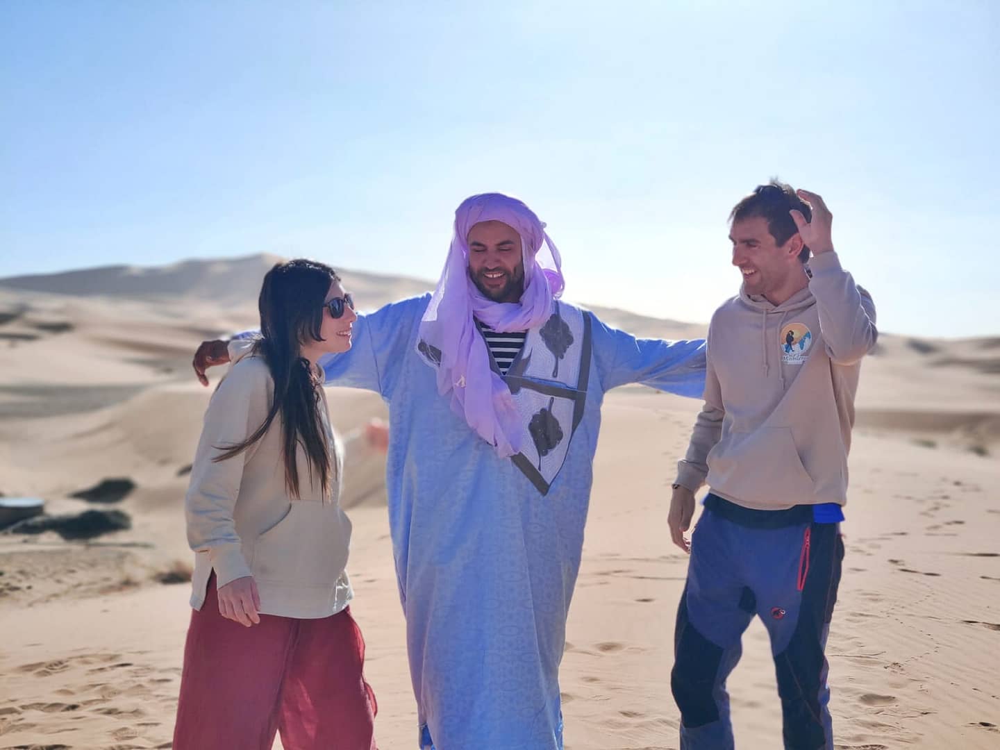 A cheerful trio walking together, with a man in traditional Moroccan attire between two tourists in the desert.