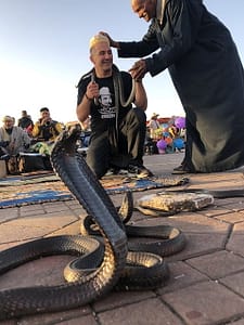 Man in traditional Moroccan attire performing a cobra snake charming act in Marrakech's bustling Jemaa el-Fnaa Square.