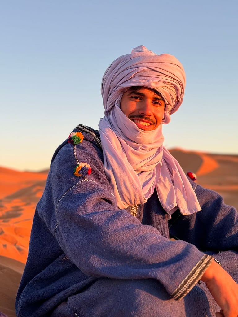 Nomad sitting on top of dune with a nice smileA smiling Berber man wearing a traditional blue turban adorned with colorful pom-poms in the Sahara Desert at sunset.